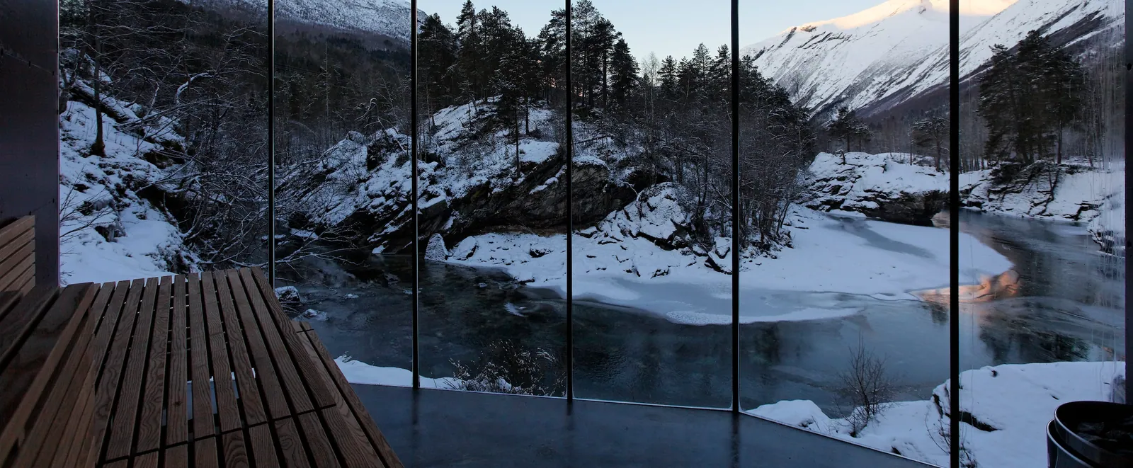 a wooden sauna bench and heater before a floor-to-ceiling glass wall over a snowy river gorge