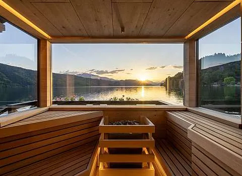 timber sauna benches and stove facing a full-width window onto the lake at sunset