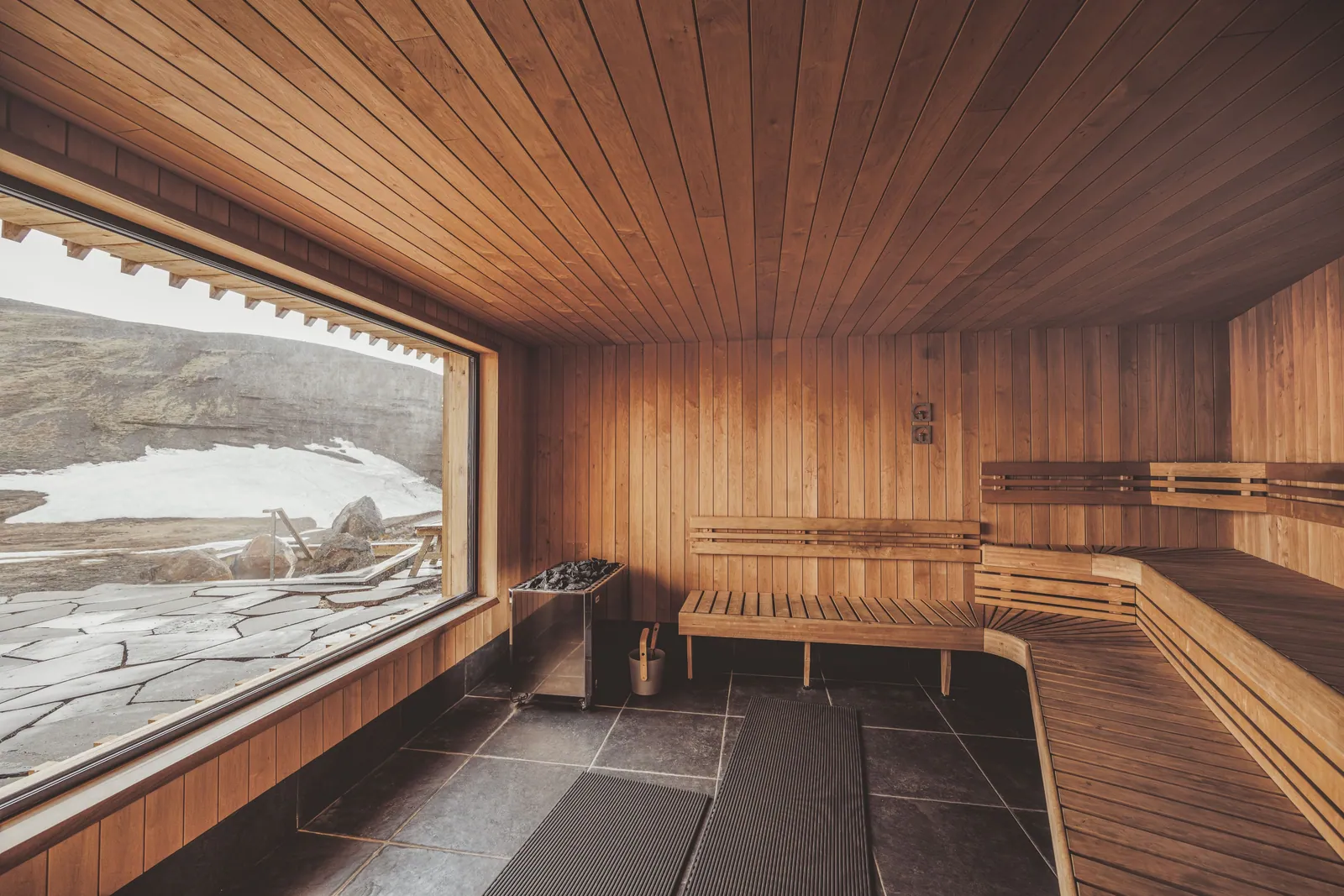 a wood-lined sauna with l-shaped benches and a big window onto snow patches and rocky slopes