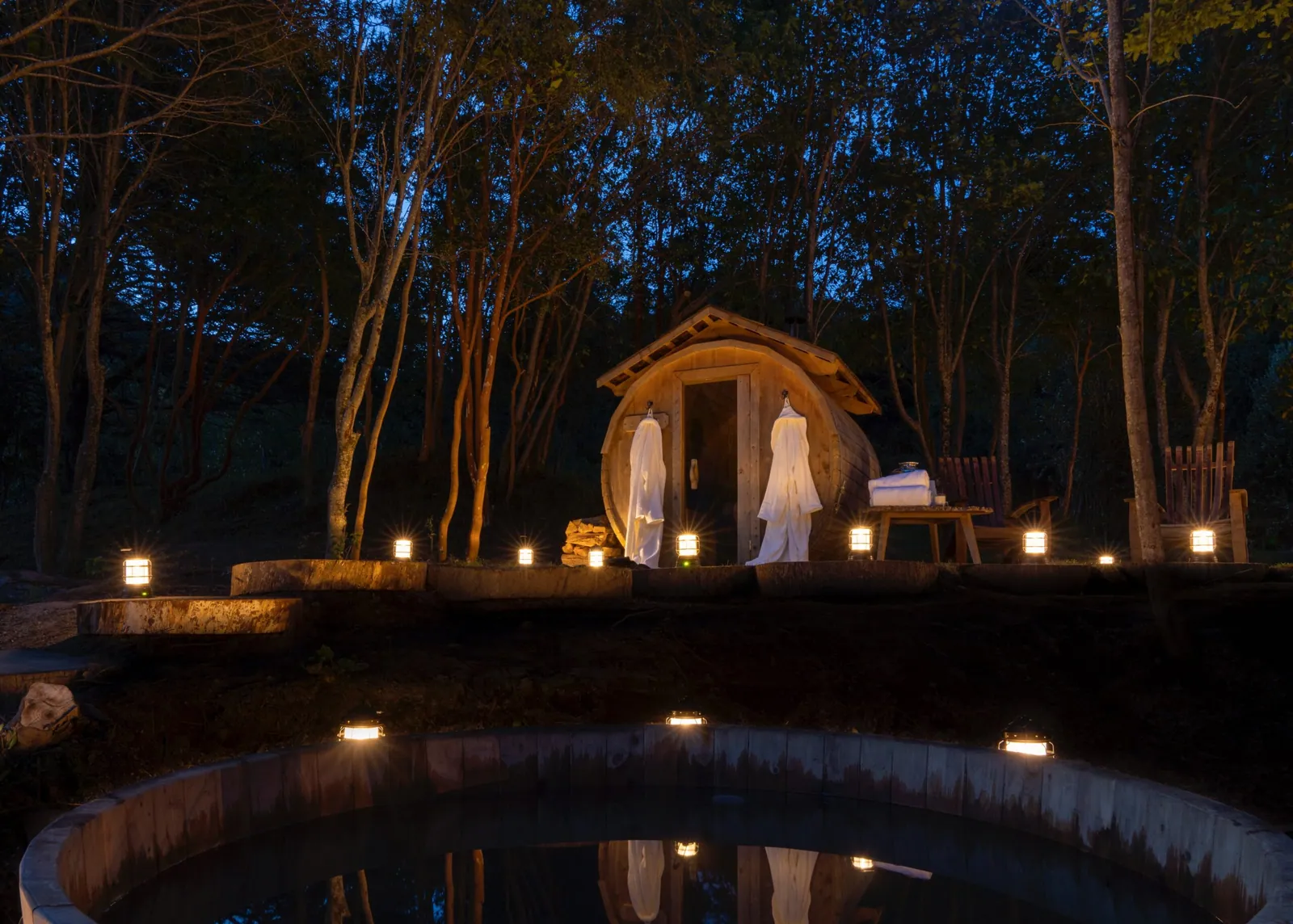 a wooden sauna cabin by a pond at dusk, lit with lanterns