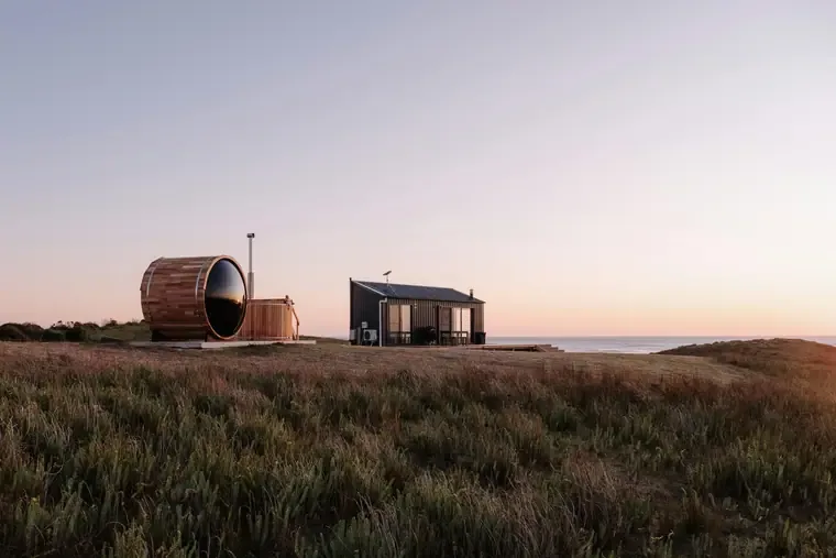 a cedar barrel sauna with a glass viewing dome and a hot tub beside a hut on a coastal headland at dusk